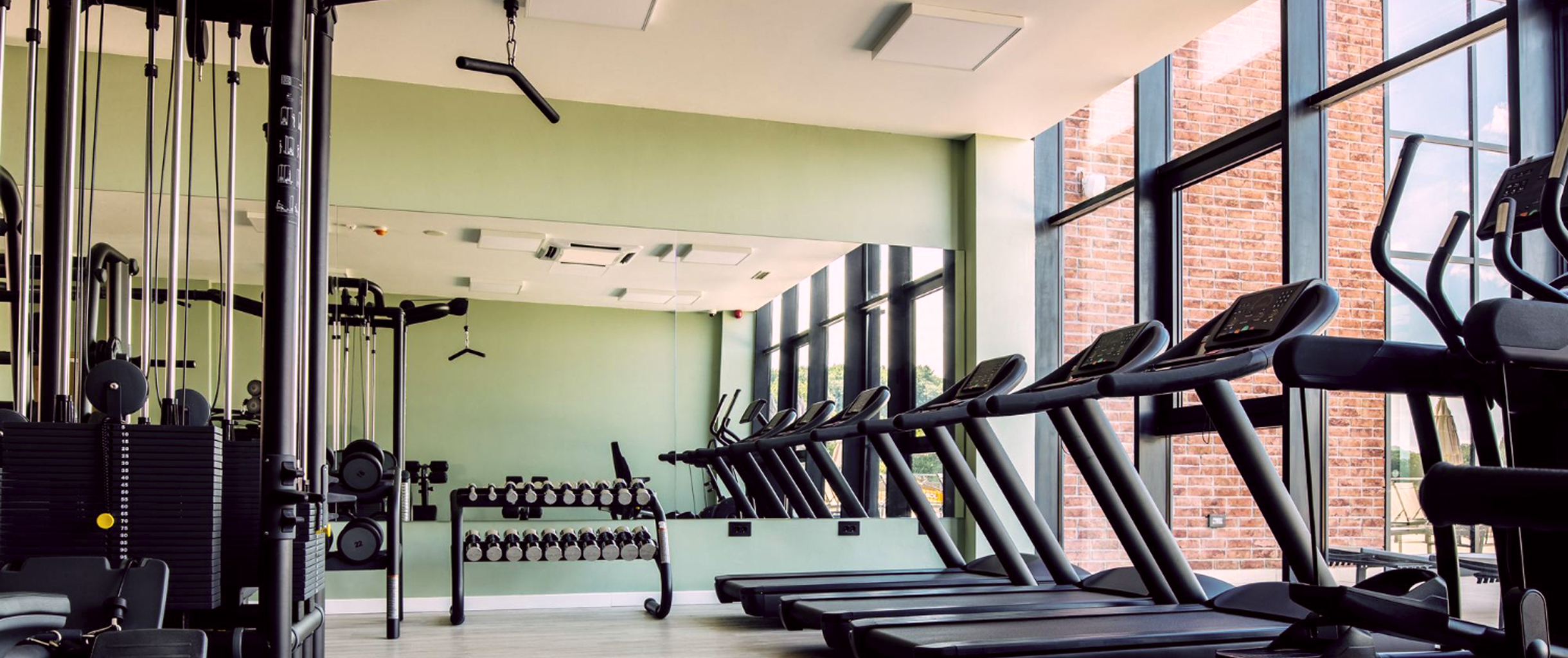 тренажерный зал с панорамными окнами. Справа находятся беговые дорожки, впереди ряд гантелей, а слева различные тренажеры